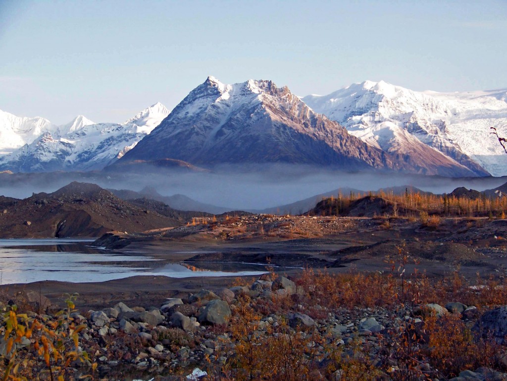 snowy mountains behind fall-colored foliage, rocks and the water